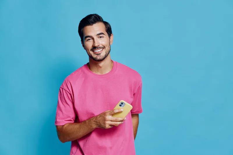 Smiling happy cheerful young man wearing pink t-shirt looks at the camera while holding a mobile phone in his hand against a vibrant blue background.