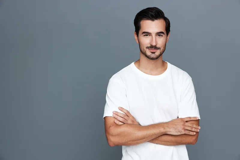 Confident young man wearing a casual white t-shirt stands with arms crossed, smiling at the camera against a neutral grey background. Professional portrait concept.
