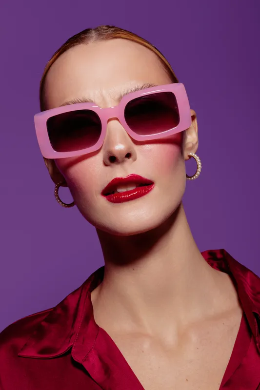 Close-up portrait of a fashionable woman wearing large pink sunglasses and red lipstick against purple background. Concept of style, beauty, makeup, and modern fashion accessories.