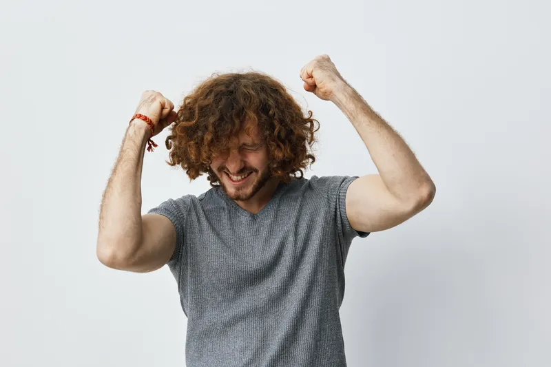 Happy man celebrating victory with curly hair in casual grey shirt against light background, showcasing emotions of joy and excitement