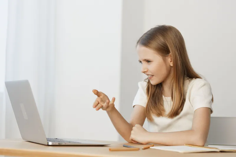 Young girl engaging in a lively online conversation at her desk, showcasing emotions of enthusiasm and curiosity, dressed in a white shirt