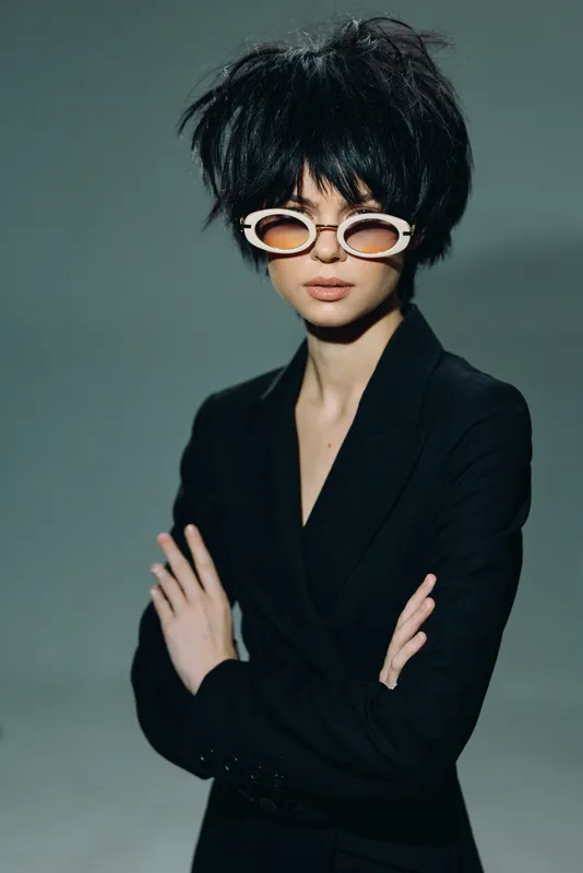 Stylish woman with short black hair wearing white sunglasses and a black blazer, posing with crossed arms against a gray background.