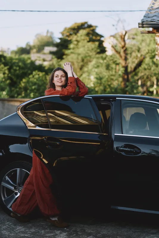 Cheerful woman outdoors in a red dress stepping from a sleek car, sunlit moment, relaxed pose, fresh breeze, vibrant colors, lush garden backdrop