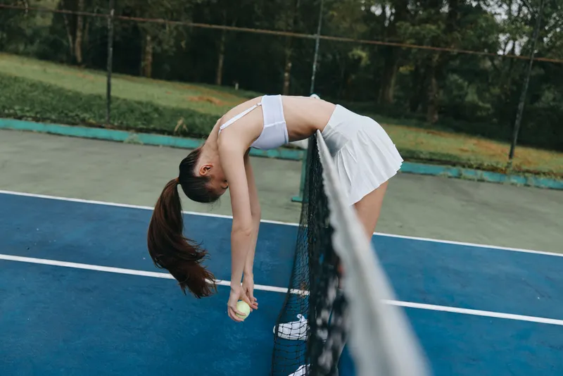 A young female tennis player in a white skirt and sports top resting against the net, showcasing emotions of fatigue and determination on a sunny day