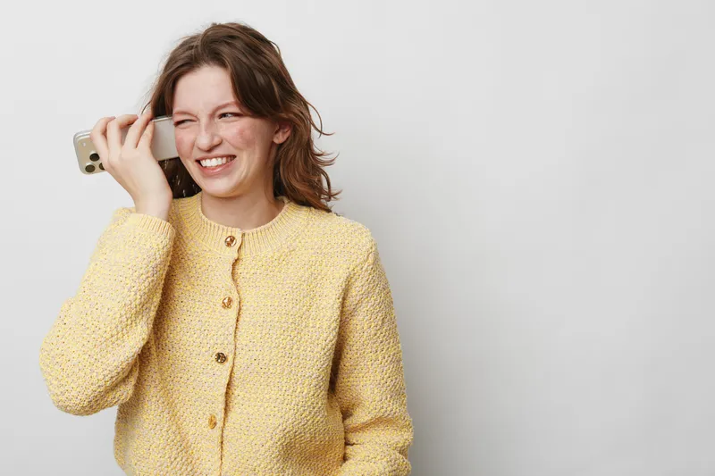 Smiling young woman in yellow knitted sweater holds smartphone near ear, laughing during phone call. Casual lifestyle, communication, happiness, and positive emotions on neutral background.