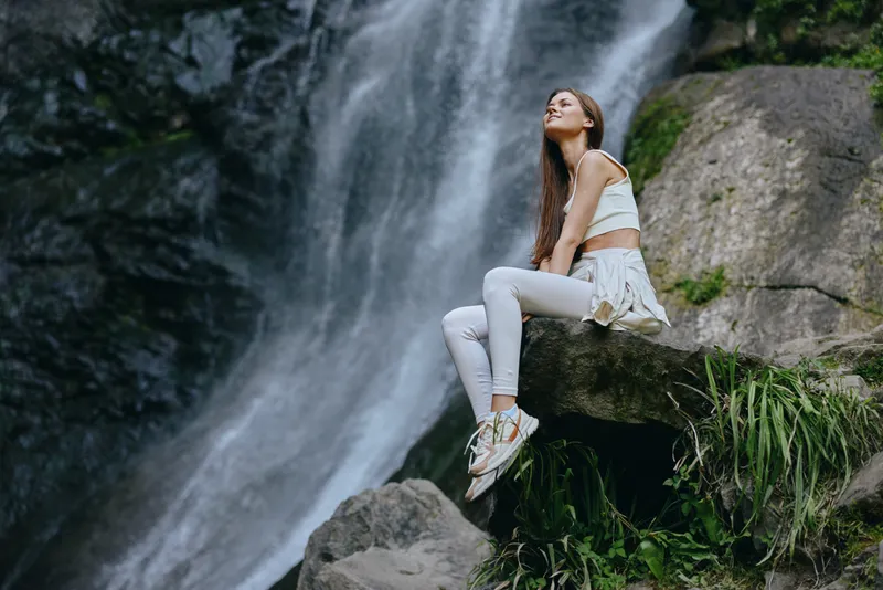 Young woman sitting on rock near waterfall in nature, wearing casual white outfit, enjoying peaceful outdoor moment with relaxed expression and natural background.