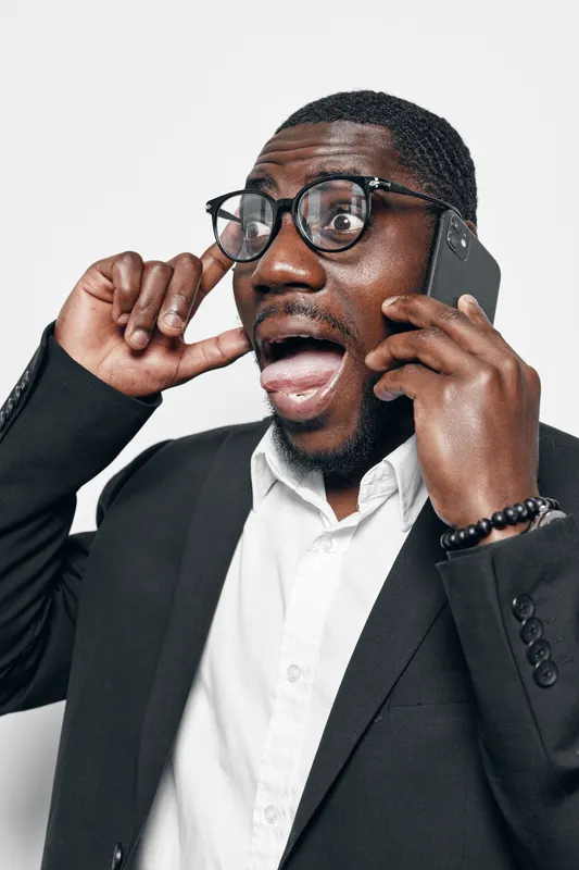 Black man with dark skin wearing glasses and black suit talking on smartphone with surprised expression. African ethnicity, communication, technology, emotion, business, portrait, casual.