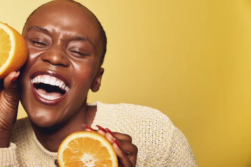 Happy African woman with dark skin laughs joyfully while holding two halves of fresh orange. Close-up on yellow background. Concept for health, freshness, and natural beauty.