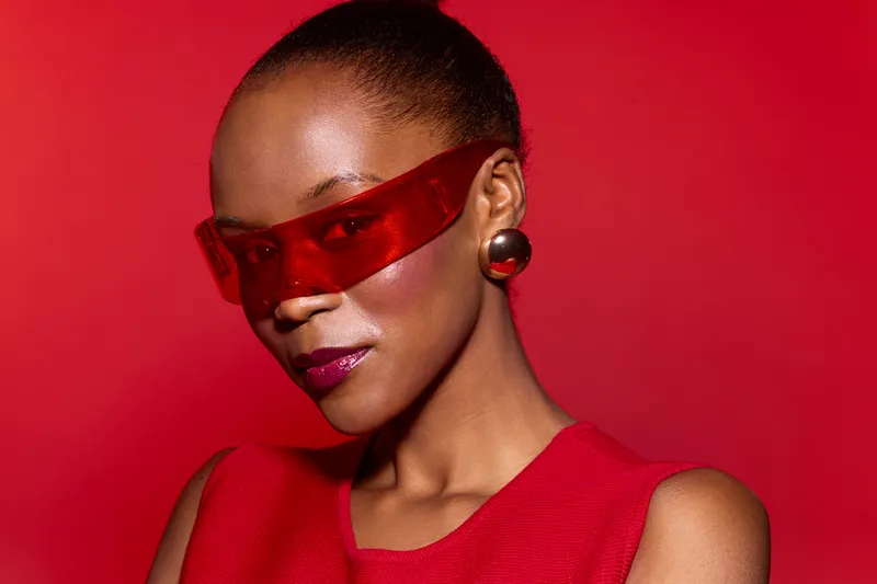Portrait of a confident black woman with dark skin wearing red sunglasses and red sleeveless top against red background. Fashion concept with bold color and modern style.