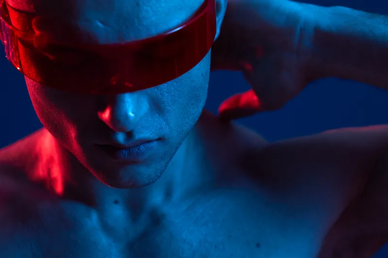 Stylish man with futuristic sunglasses showcasing a unique fashion statement in a vibrant blue and red light setting. Ideal for technology and fashion themes.