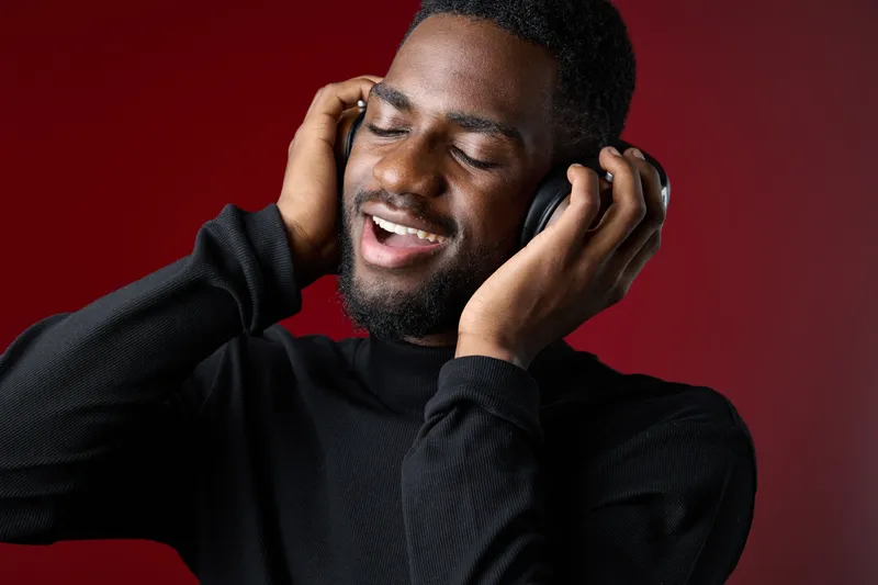 Joyful young Black man enjoying music with headphones, smiling against a deep red background, conveying happiness and relaxation through music
