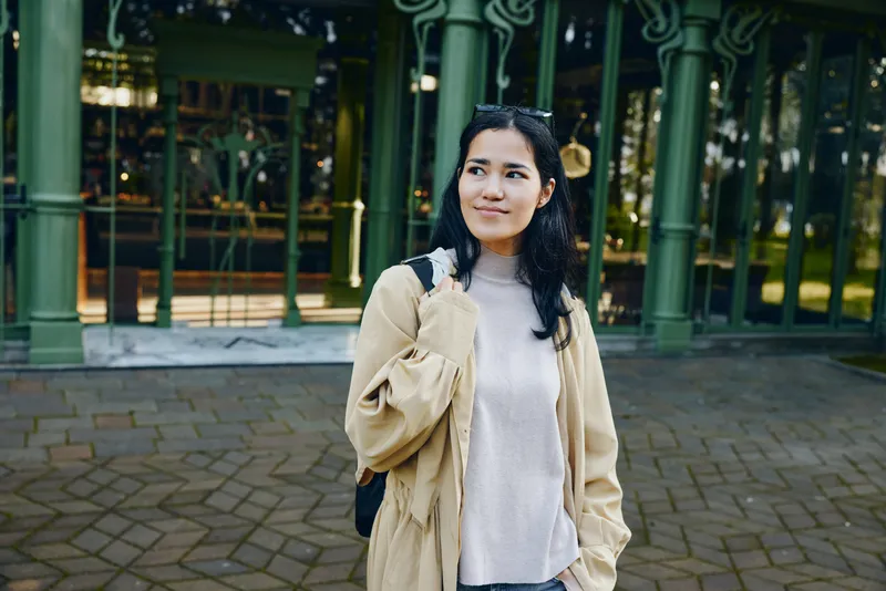 Woman in a light coat outdoors on a city street, carrying a backpack, smiling as she explores urban scenery, daylight, casual fashion and travel vibe, her beige jacket blending with architecture for