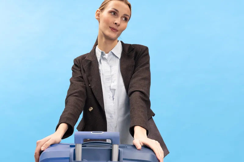 A thoughtful woman in a brown coat holds a blue suitcase against a blue background. Concept of travel, decision making, and planning for a trip or journey.