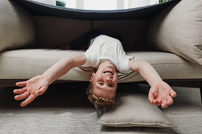 Happy child lies upside down on couch with arms outstretched, smiling joyfully in cozy living room. Concept of playful childhood and carefree moments.