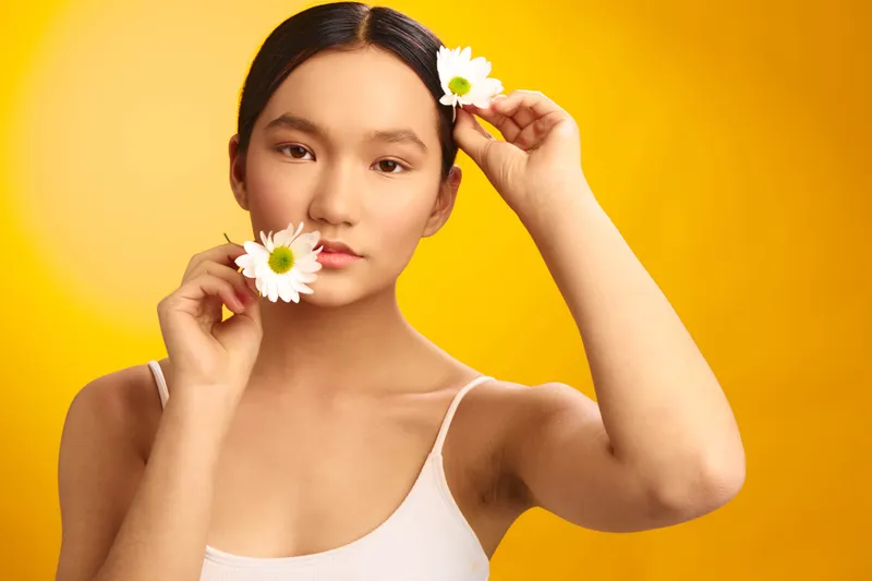 Asian woman stands against a bright yellow backdrop, gently holding white daisies near her face, creating a soft, fresh beauty portrait with a calm and natural vibe.