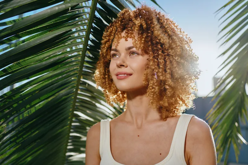smiling woman with curly hair enjoys sunny tropical background, natural light highlights her clear skin and relaxed expression in warm outdoor setting