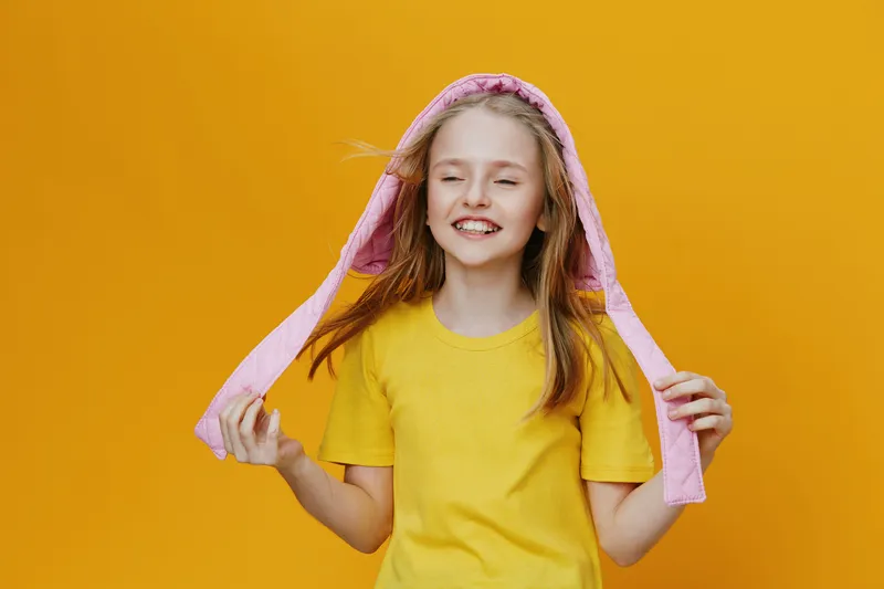 Cheerful girl in yellow shirt with pink hoodie, smiling against a bright yellow background, embodying joy and youthfulness