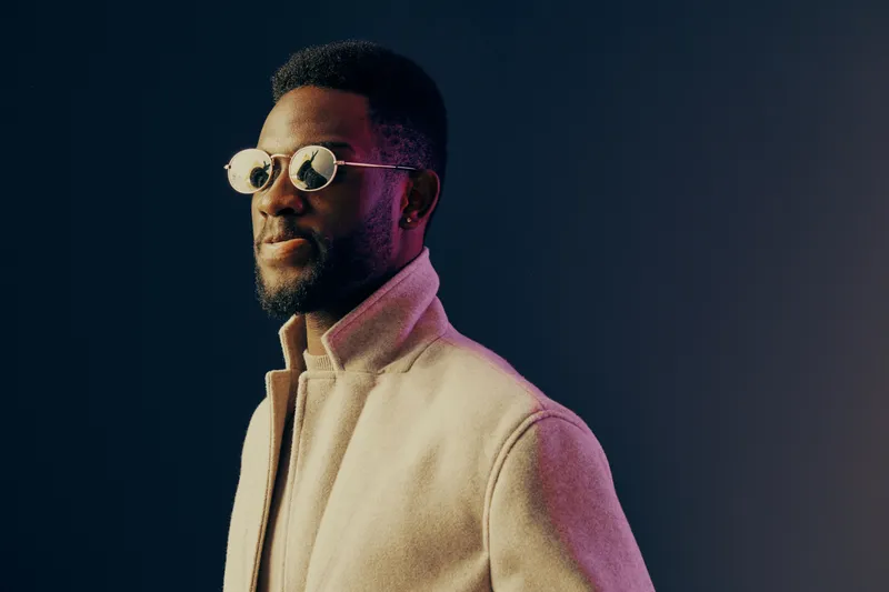 Stylish man in sunglasses wearing a light coat, showcasing confidence against a dark gradient background suitable for fashion promotions or lifestyle branding