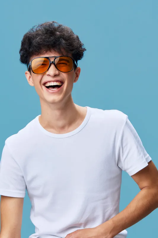 young man laughing wearing orange sunglasses and white t-shirt against blue background, joyful expression and casual style bright cheerful mood