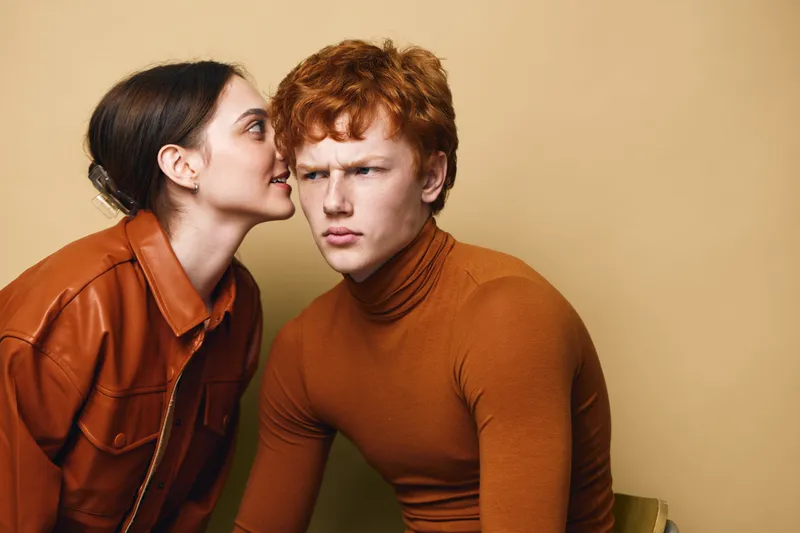Young woman whispering secret to puzzled man in brown sweater and leather jacket on beige background. Conversation, communication and facial expressions with closeup portrait mood.