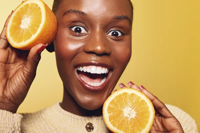 Happy African woman with dark skin holding two orange halves near her face on yellow background. Concept of healthy lifestyle, fresh fruit, natural vitamin C, and joyful wellness.