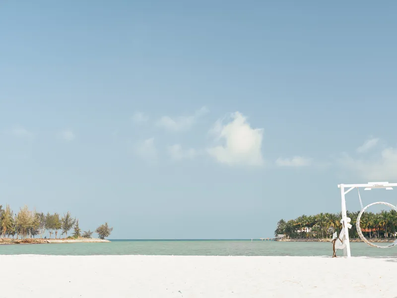 A tranquil beach scene shows a lone figure near a white circular frame on powdery sand, with calm turquoise water and a bright blue sky, evoking serenity.