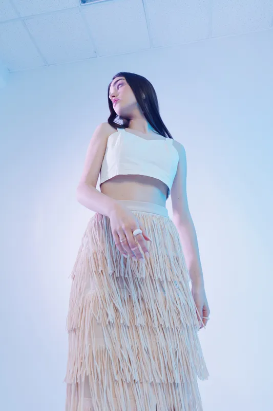 Young woman in a white crop top and fringed skirt poses confidently against a bright ceiling background. Concept of woman beauty fashion and modern style.
