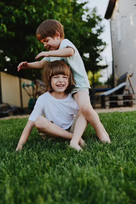 Happy kids playing outdoors on green grass, one boy sitting while the other stands behind him, enjoying a sunny day filled with laughter and joy.
