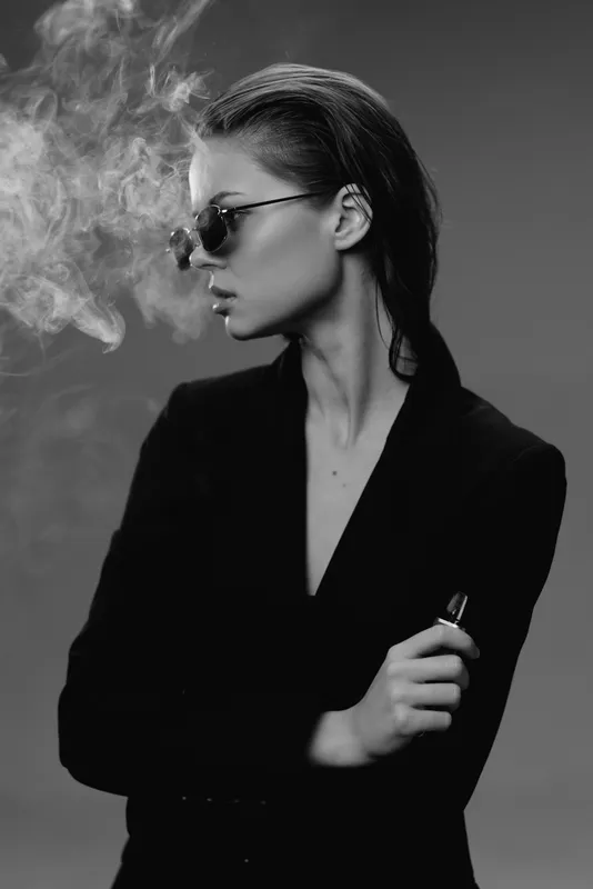 Black and white portrait of a stylish woman with sunglasses vaping, exhaling smoke, wearing a dark blazer