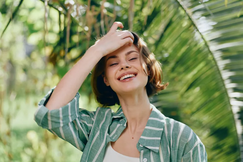 Cheerful woman outdoors in sunlit light, smiling with hand on her forehead, wearing a casual striped shirt.