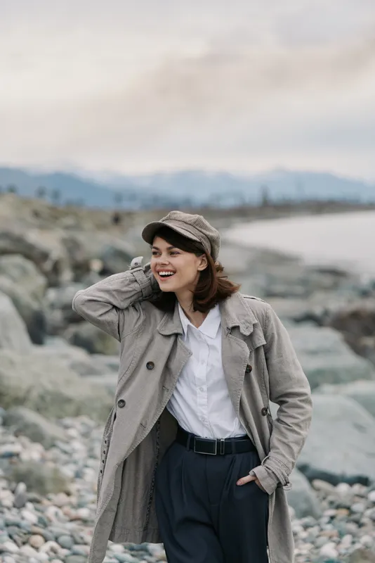Smiling woman wearing a knitted coat and cap stands on rocky shore near water, enjoying outdoor autumn day. Casual style with natural background and relaxed happy mood.