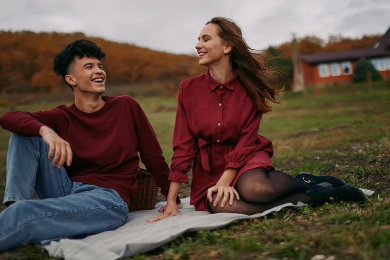 Candid outdoor portrait of a genuine young couple sharing a relaxed picnic moment on a blanket, natural smiles and honest interaction capturing real connection and warmth.
