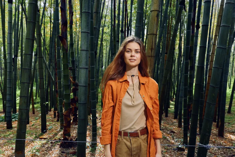 Woman bamboo forest portrait nature standing fashion outdoors: Young woman with long brown hair in orange coat stands among tall bamboo stalks, relaxed serene face in soft sunlight.