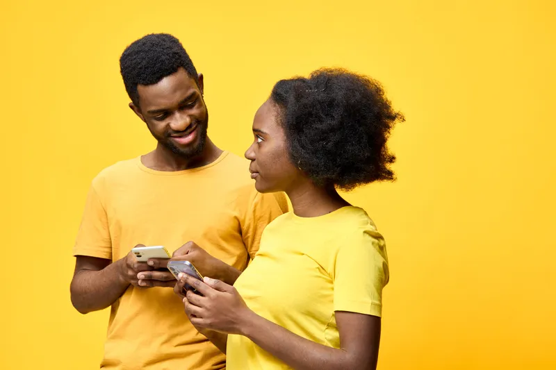 Happy young African couple with smartphones, wearing matching yellow shirts, enjoying communication against a bright yellow background, perfect for social media and relationships