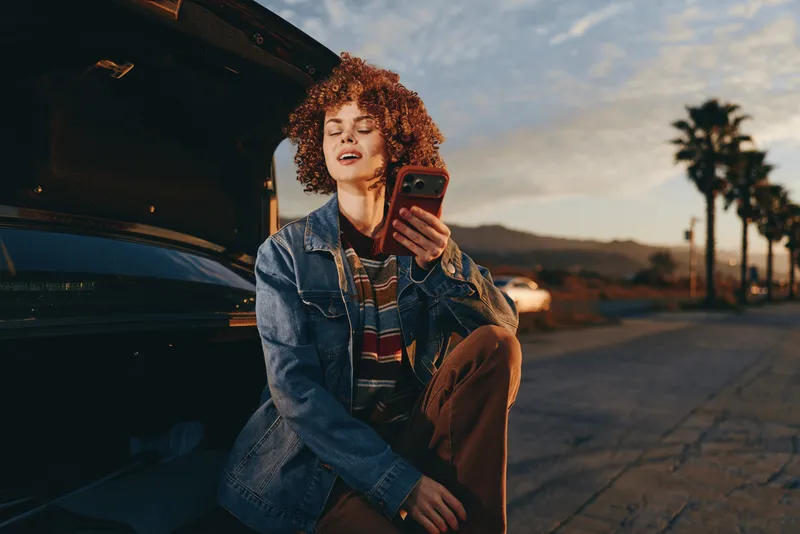 Woman lifestyle portrait outdoors by parked car at sunset curly haired in denim jacket holding phone for selfie relaxed smile street pavement palm trees golden evening light