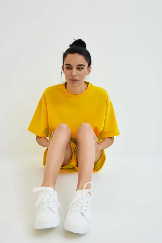 Young woman sitting on the floor wearing bright yellow casual dress and white sneakers, with natural makeup and hair tied in a bun against a plain white background.