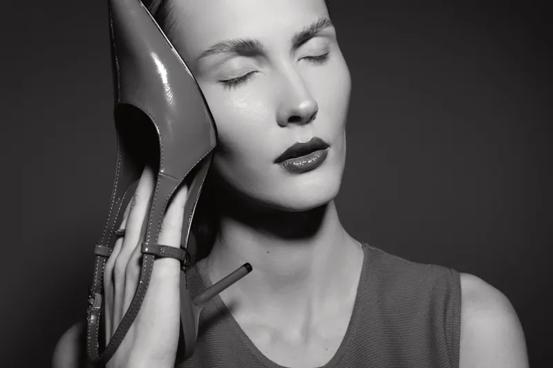 White woman with pale skin holds high heel shoe to her ear with closed eyes in black and white photo.