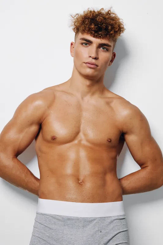 Young strong muscular man with curly hair and tanned skin standing shirtless in undergarments against white wall. Fitness model reflecting health and body confidence action.