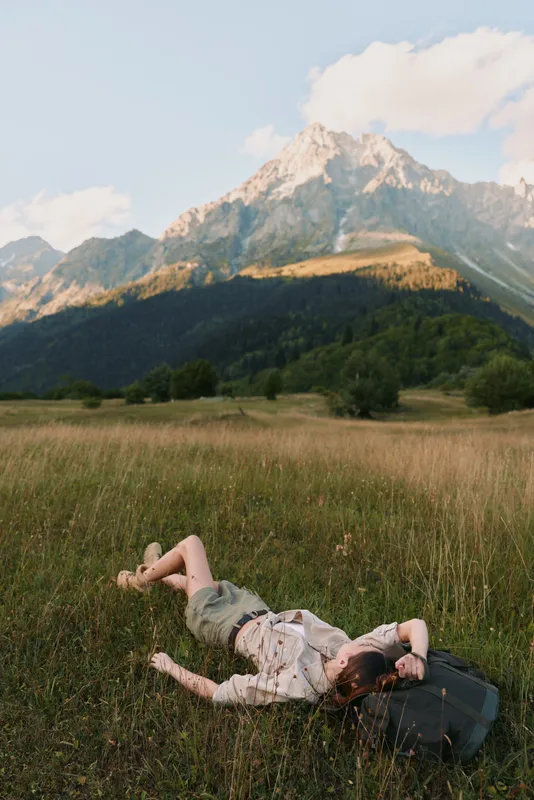 A woman rests in a sunlit meadow with a backpack beside her, mountains rising in the distance. A calm travel scene conveying rest, exploration, and connection with nature.