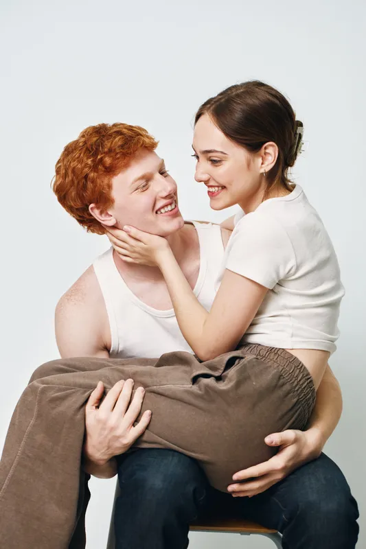 Young couple in casual clothes smiling and embracing, man carrying woman on lap in cozy indoors with white background and affectionate happy mood in studio.