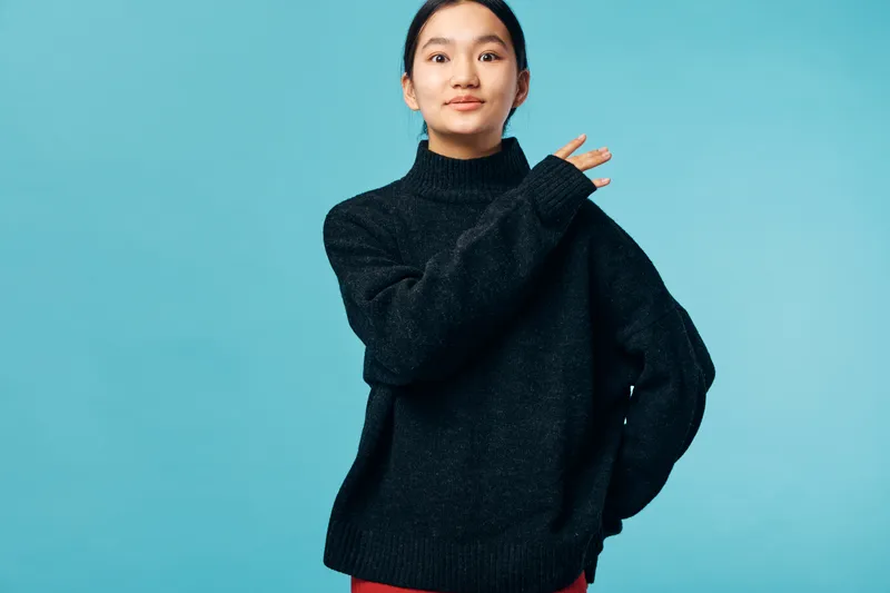 Young woman wearing black sweater posing with confident expression against blue background. Female model with casual style in studio lighting, portrait with hand gesture and relaxed mood.
