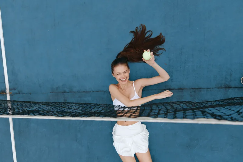 A cheerful young woman of Asian descent plays tennis with a smile, wearing sports attire, set against a vibrant blue background that enhances the energetic mood