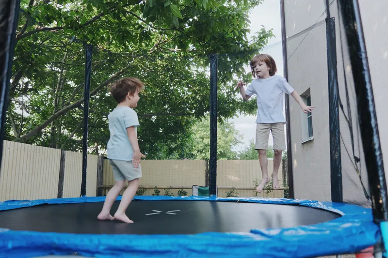 Two playful boys joyfully jumping on a trampoline, showcasing a carefree childhood spirit in a vibrant outdoor setting surrounded by lush greenery.