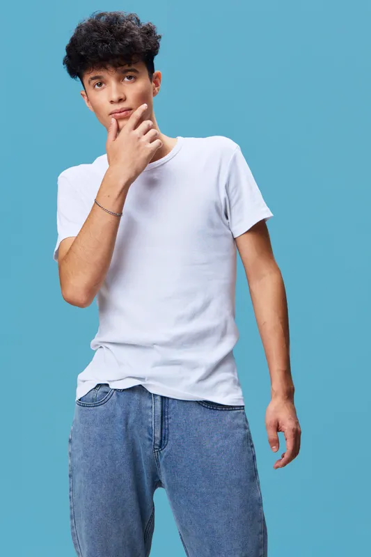 young man in white t-shirt and jeans posing thoughtfully against blue background, casual style with curly hair and relaxed posture