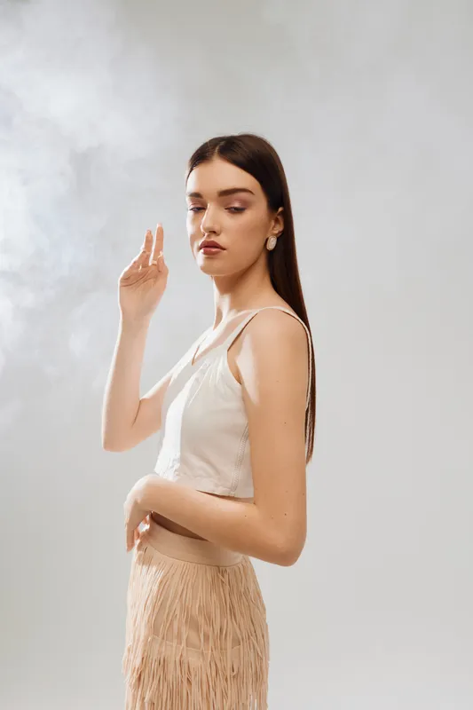 Young woman with long dark hair poses in a beige top and fringed skirt against a neutral background. Concept of beauty, fashion, elegance, and modern style in studio lighting.