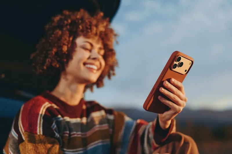Smiling woman lifestyle takes smartphone selfie outdoors, curly hair visible, striped shirt, warm sunset sky, distant landscape, candid travel moment, relaxed fun