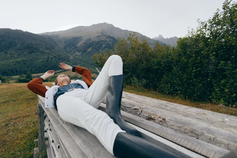 Woman relax on wooden bench in countryside meadow with mountains in background, casual outfit and rubber boots, outdoor leisure and peaceful nature escape in scenic rural landscape.