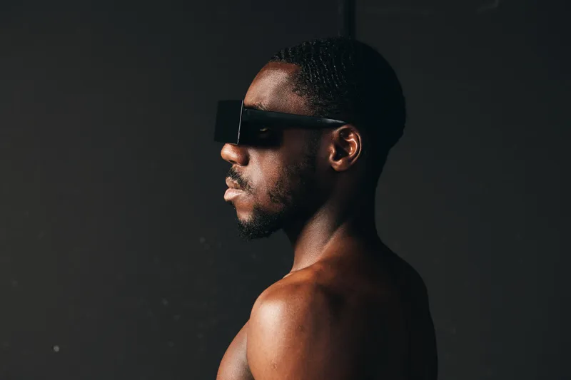 Creative visual designer style portrait of black African man with dark skin wearing futuristic black sunglasses in profile on dark background with dramatic lighting.