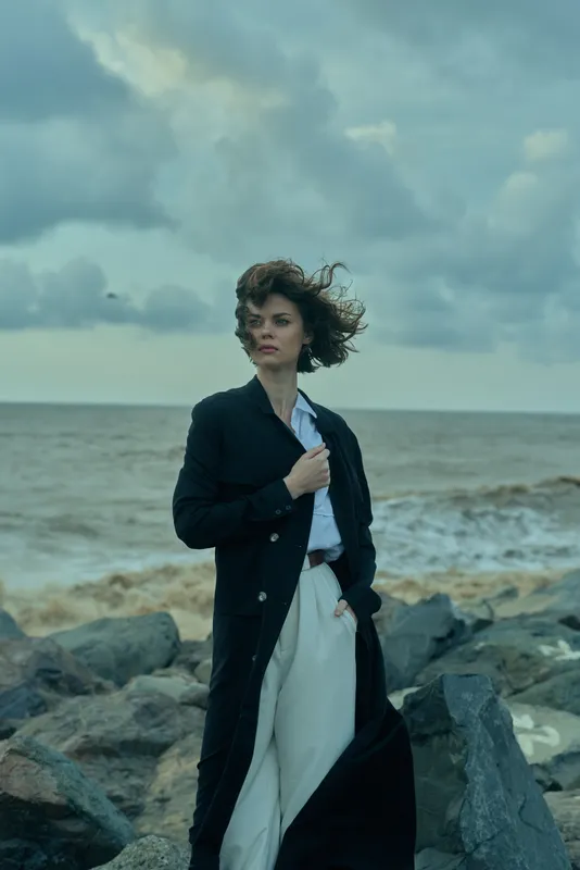Woman in a long dark coat stands on a rocky shoreline as wind sweeps her hair, navy suit and light pants, moody sea backdrop and dramatic skies
