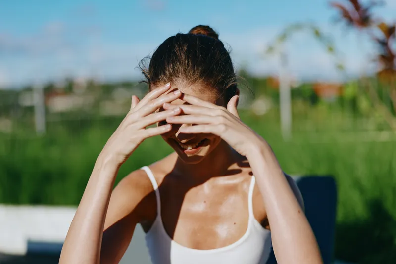 Young woman in white tank top shielding her face from the sun with hands outdoors in a sunny garden setting. Concept of summer lifestyle, wellness, and natural beauty.